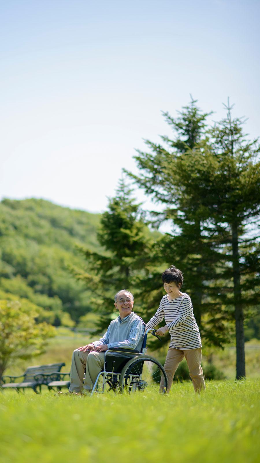 晴天の草原で車椅子に乗った高齢者を介護する様子の写真