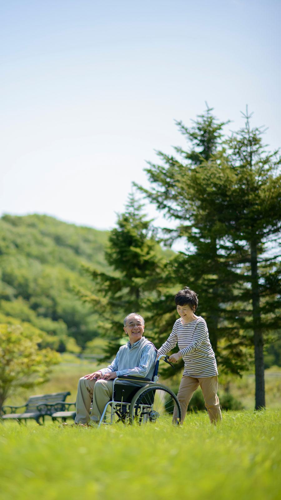 晴天の草原で車椅子に乗った高齢者を介護する様子の写真